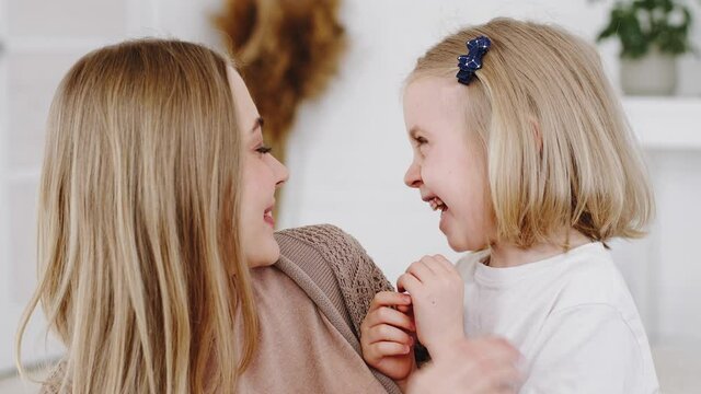 Portrait of blonde caucasian woman happy mother nanny babysitter hugs little daughter baby cute girl tickles cuddles kisses on cheek child play game at home, kid laughs sincerely loudly from tickling