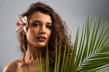Pretty female model with naked shoulders and curly brown hair looking at camera and holding palm leaf. Young woman with perfect fresh makeup posing with pink orchid flower behind ear isolated on gray.
