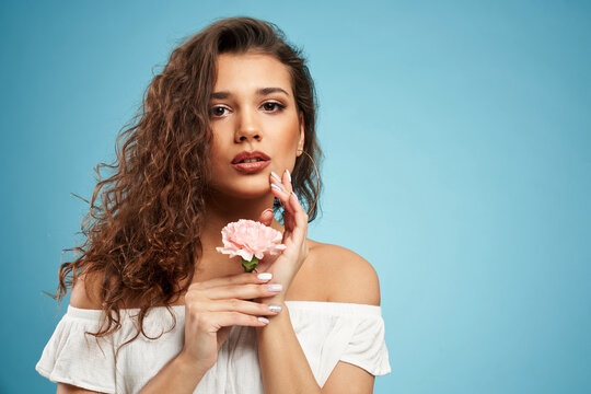 Stunning brunette female model with curly hair looking at camera and touching face, mouth open, isolated on blue. Portrait of pretty young woman with perfect makeup posing with pink flower in hand.