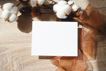 Blank white card on wooden background with cotton flower, dry thistle and organza ribbon. Elegant feminine composition. Empty space for text. Copy space.
