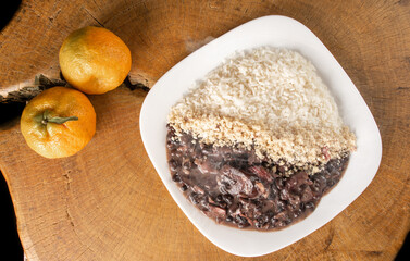 Brazilian feijoada, white dish with a delicious Brazilian feijoada, rice and farofa, with two oranges on rustic wood, top view.