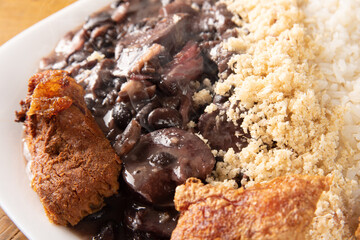 Brazilian feijoada, white dish with a delicious Brazilian feijoada, rice, farofa, and fried pork with two oranges on rustic wood, selective focus.