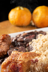 Brazilian feijoada, white dish with a delicious Brazilian feijoada, rice, farofa, and fried pork with two oranges on rustic wood, selective focus.