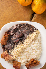 Fototapeta na wymiar Brazilian feijoada, white dish with a delicious Brazilian feijoada, rice, farofa, and fried pork with two oranges on rustic wood, top view