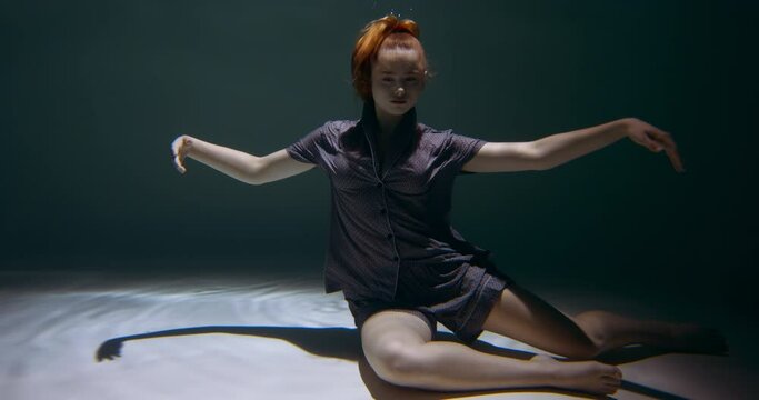 Young beautiful peaceful redhead woman wearing night pyjamas sits down deep under water on dark pool floor slow motion.