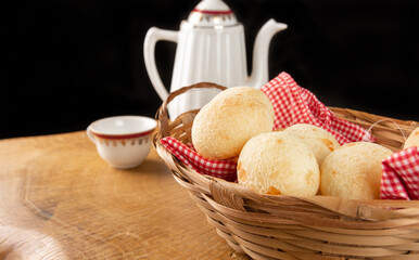 Brazilian cheese bread, checkered cloth basket with cheese breads on rustic wood with teapot and cup of coffee in the background, selective focus.