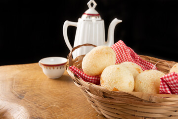 Brazilian cheese bread, checkered cloth basket with cheese breads on rustic wood with teapot and cup of coffee in the background, selective focus.