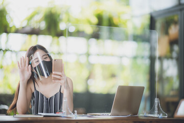 young Asian business woman working in cafe with medical face mask and table shield protection, social distancing of the new normal lifestyle after epidemic of coronavirus COVID-19