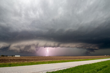Lightning strikes as a supercell thunderstorm approaches a rural road. A shelf cloud marks the edge of the storm.