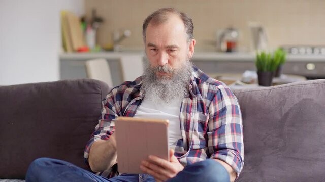 Joyful senior man with gray beard having fun playing video game on tablet computer sitting cross-legged on sofa at home