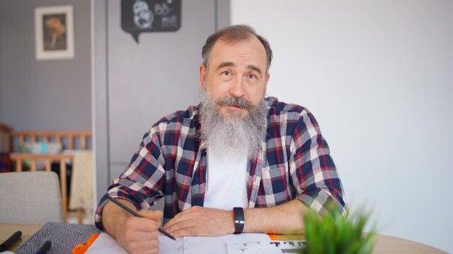 POV shot of senior man with gray beard speaking and taking notes while having online consultation, class or business meeting from home during coronavirus stay at home order