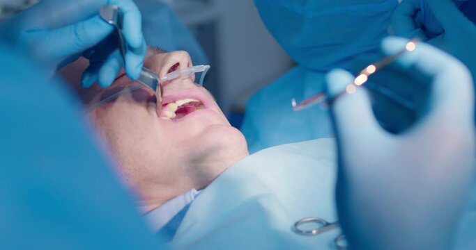 Close-up an adult male patient is under anesthesia while the dentist is holding a dental retractor and pulling forceps with the surgical thread and a nurse is inserting a saliva ejector.