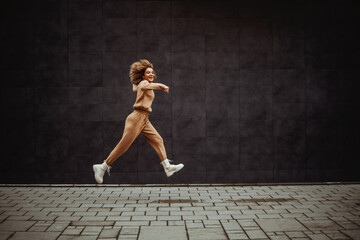 Smiling trap girl in tracksuit running next to a gray wall. - obrazy, fototapety, plakaty