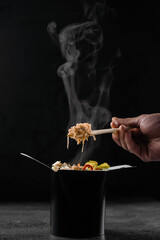 Steaming hot rice in wok box on black background. Holding rice with chinese chopsticks. Asian spicy dish for street restaurant and fast food delivery service.