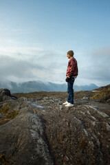 Naklejka na ściany i meble Hombre joven parado en la cima de una montaña mirando el paisaje con la nubes azules de fondo