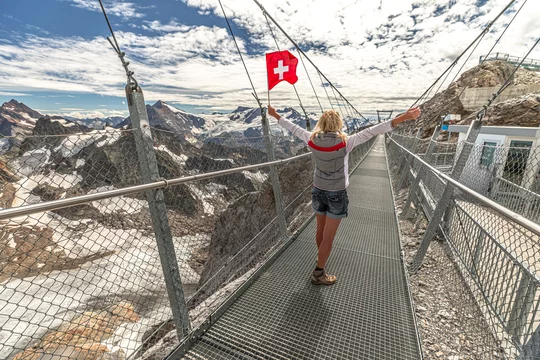 Woman with Swiss flag on Titlis cliff walk suspension bridge. Top of Titlis  mountain with glacier in the Uri Alps. Viewpoint 3028 m in cantons of  Obwalden and Bern, Switzerland, Europe. Summer