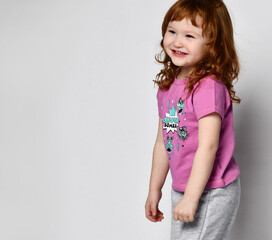 Shy smiling little girl smiling cutie baby in sportswear studio. Little adorable girl laughs out loud. A child in a pink T-shirt with a cute fairy print and sweatpants. T-shirt lettering: girl bomb