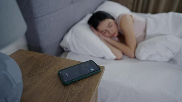 A cute young girl is sleeping in a white bed and is woken up by alarm on the phone, she doesn't want to pick up and continues to sleep