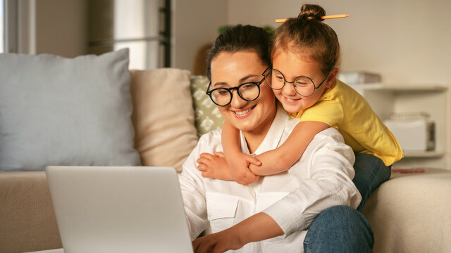 Young happy woman mother in glasses working on laptop with little girl daughter wearing glasses, excited child hugging smiling mom while learning online together in light cozy living room at home