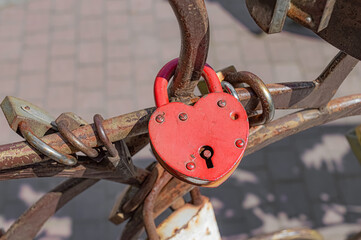 Red closed heart-shaped padlock hanging near other padlocks on old decorative metal fence
