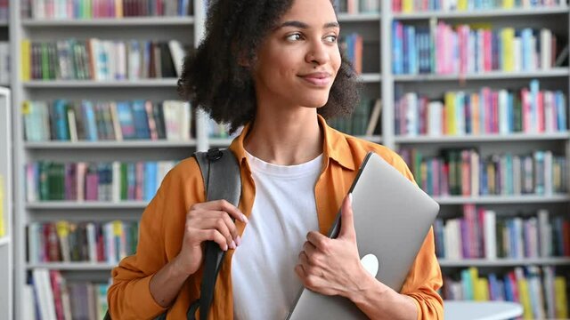 Confident happy attractive teenage african american female student, goes to the camera, looks to the side then to the camera, in campus library, holds laptop and backpack, friendly smiles