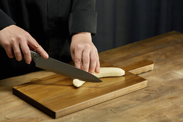 A woman slices a banana with a large knife on a wooden chopping board on the kitchen table. Slicing by knife on wooden table dark background