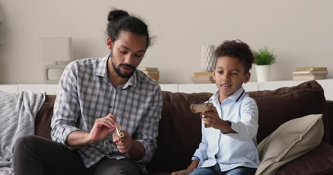 Loving African dad play toy guns with preschool adorable son. Family sit on sofa communicating enjoy playtime with modern playthings, father teach kid on weekend leisure at home. Boyish hobby concept