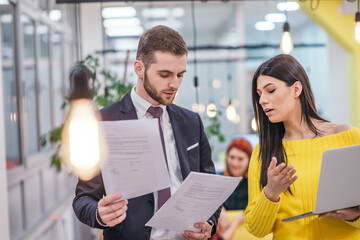 Businessman boss checking the paperwork handed over bus his female employee, while standing in a modern startup office.