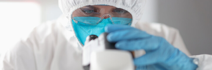 Doctor in protective suit and glasses looks through microscope