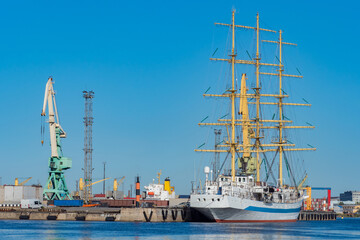 Fototapeta premium The yacht is moored at the pier in the port. Sailboat close-up. A ship with sails on the background of the port and the blue sky. Navigation and travel.