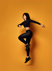Fototapeta na wymiar Young smiling sexy pretty lady in black sportswear jumping during workout over yellow background, side view. Beautiful women and active sport lifestyle concept