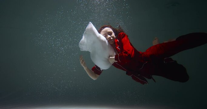 Cinematic shot, young beautiful mixed race woman in red pyjamas sinks under water to fall asleep with pillow slow motion
