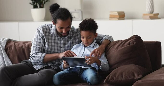 Young African loving dad teach son to use digital tablet explain new program develop preschool kid, learn educational application seated on sofa at home. New generation and modern tech overuse concept