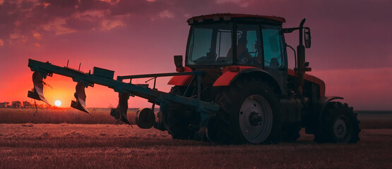 Tractor with a raised plow in an agricultural field against the backdrop of a sunset. Plowing after harvest. Concept, agriculture, food industry. Selective focus, toned photograph. - obrazy, fototapety, plakaty