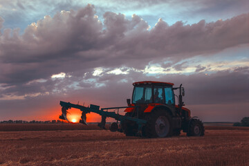 Obraz premium Tractor with a raised plow in an agricultural field against the backdrop of a sunset. Plowing after harvest. Concept, agriculture, food industry. Selective focus, toned photograph.