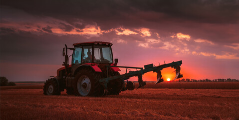 Tractor with a raised plow in an agricultural field against the backdrop of a sunset. Plowing after harvest. Concept, agriculture, food industry. Selective focus, toned photograph. © Vladimir Kayukov