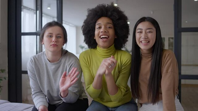 Video call POV of three cheerful multiethnic women waving and smiling at webcamera. Portrait of positive multinational female friends chatting online from living room indoors. Slow motion