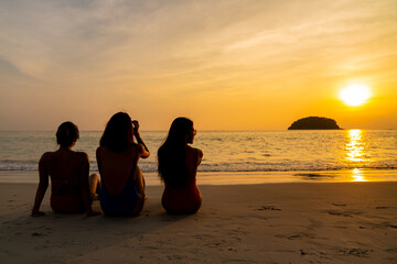 Group of Asian woman friendship in colorful swimwear sitting on the beach and talking together at summer sunset. Female girl friends enjoy and having fun outdoor activity lifestyle on summer vacation