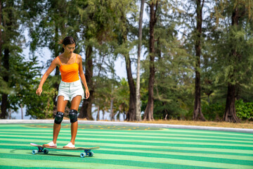 Attractive Asian woman with safety skateboarding knee pad skating at skateboard park by the beach. Happy female enjoy summer outdoor active lifestyle play extreme sport surf skate at public park.