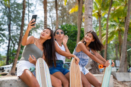 Happy Asian woman girl friends sit on the beach after skateboarding and use smartphone taking selfie together. Female friendship enjoy and having fun outdoor activity lifestyle in summer vacation.