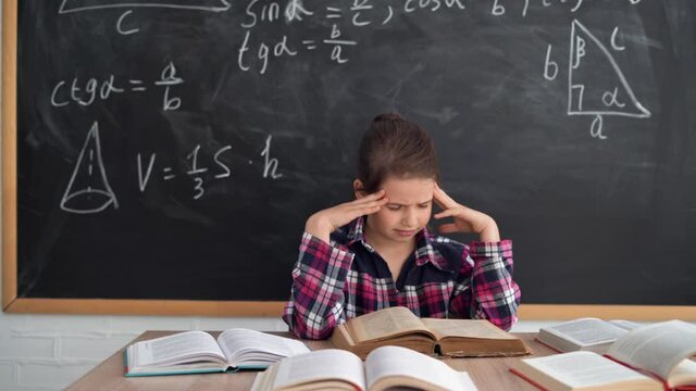A Caucasian schoolgirl girl sits at a table against the background of a chalk board. massage the weights with your fingers. The concept of overwork during training.