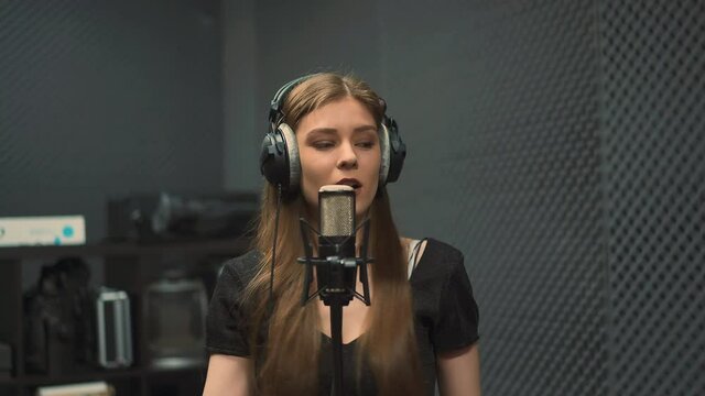 Expressive young woman singer with black headphones records new song with contemporary microphone dancing in dark audio studio close view