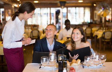 Friendly smiling female waiter consulting couple of guests choosing drinks and meals in restaurant