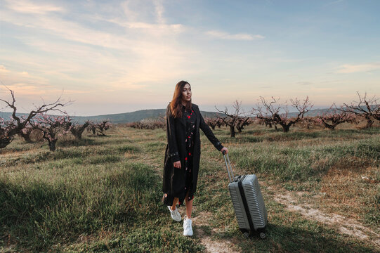 a girl traveling alone stands in a blooming garden with luggage 
