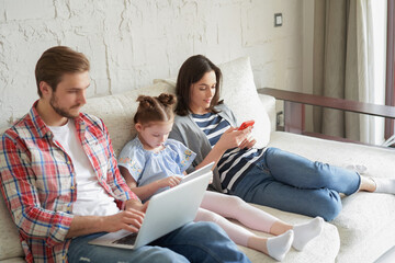 Father, mother and daughter using electronic devices sitting on sofa at living room.