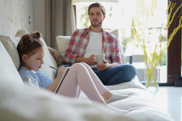 Happy father smilling daughter sitting on sofa enjoying creative activity, drawing pen pictures in albums, father and daughter spend free time together.