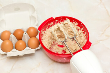 Electric beater on a red bowl with dough and egg carton