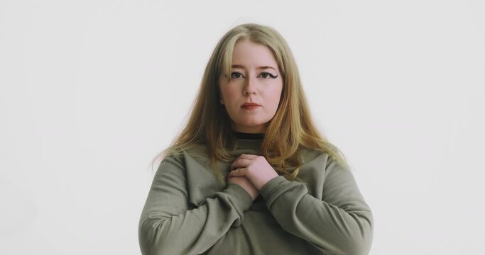 Hopeful plump blonde woman in green sweatshirt puts hands to chest posing for camera at white wall at audition closeup slow motion
