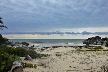 Wild beach near the ancient Mayan city of Tulum. Cloudy. The waves of the surf roll to the shore. Algae, logs on the sand. Picturesque stones by the water. Mexico