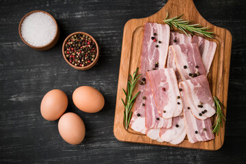 Board with raw bacon with spices, and rosemary on a black background